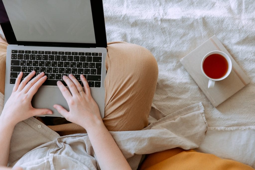 crop person using laptop with empty screen in bed near cup of tea placed on notebook
