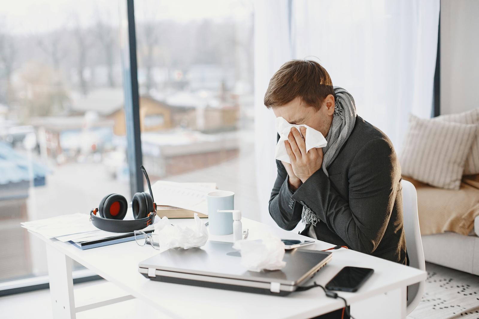 Man sneezing at home desk, indicating remote work during illness.